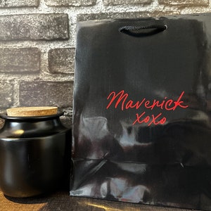 Pode incluir: Uma sacola de presente preta com um frasco de corti&ccedil;a na frente. A sacola tem o texto "Maverick xoxo" impresso em vermelho na frente.