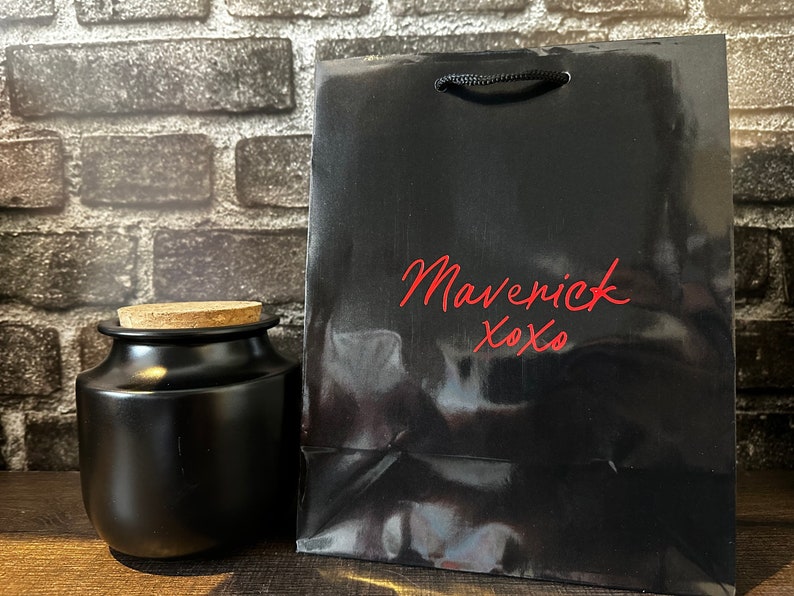 Pode incluir: Uma sacola de presente preta com um frasco de corti&ccedil;a na frente. A sacola tem o texto "Maverick xoxo" impresso em vermelho na frente.