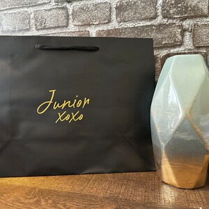 Pode incluir: Uma sacola de presente preta com uma inscri&ccedil;&atilde;o dourada que diz "Junior xoxo". A sacola est&aacute; sobre uma superf&iacute;cie de madeira ao lado de dois vasos de cer&acirc;mica geom&eacute;tricos com um esmalte azul e dourado.
