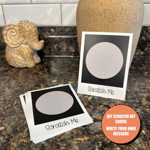 May include: A stack of white cards with black borders and a circle in the center that can be scratched off. The text "Scratch Me" is printed on the cards. The text "DIY Scratch Off Cards Write Your Own Message" is printed below the cards.