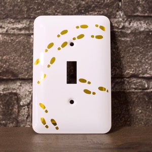 May include: White light switch cover with a gold design of footprints. The footprints are in a curved pattern, like someone is walking away.