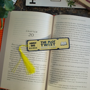 May include: A yellow bookmark with a tassel, featuring a ticket design with the text "Admit One" and "The Plot Twist Book Club". The bookmark is placed on an open book with the text "Chapter 20" visible.