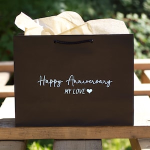 May include: A black gift bag with the words "Happy Anniversary MY LOVE" in white script. The bag has a black rope handle and is filled with cream-colored tissue paper. A white heart symbol is below the words "MY LOVE".