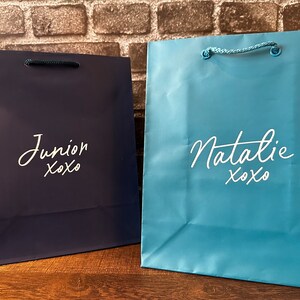Pode incluir: Duas sacolas de presente, uma azul e outra azul claro, com letras brancas. A sacola azul diz "Junior xoxo" e a sacola azul claro diz "Natalie xoxo".
