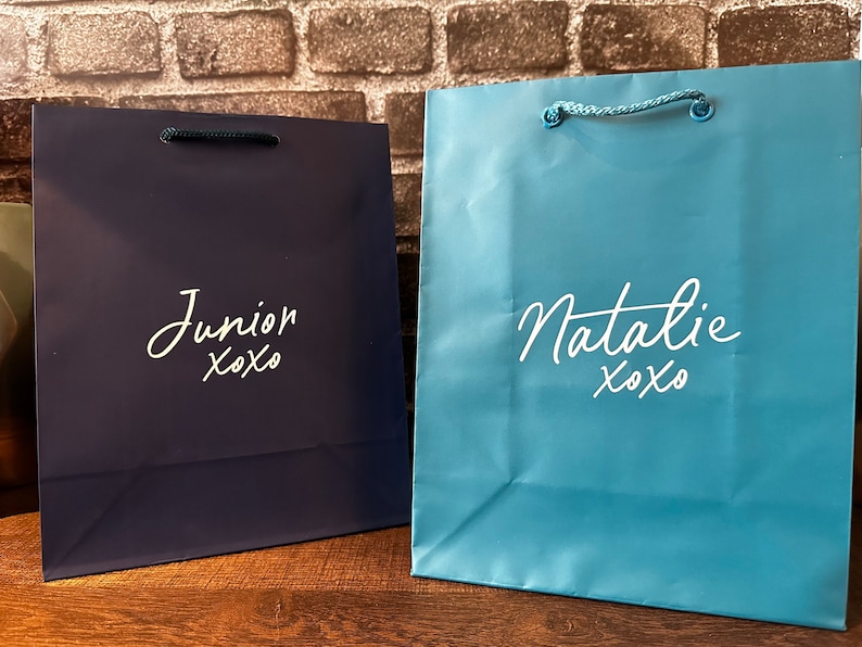 Pode incluir: Duas sacolas de presente, uma azul e outra azul claro, com letras brancas. A sacola azul diz "Junior xoxo" e a sacola azul claro diz "Natalie xoxo".