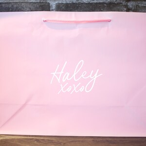 Pode incluir: Uma sacola de papel rosa com um logotipo branco que diz "Haley xoxo".