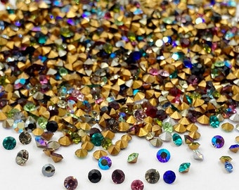 May include: A large pile of colorful rhinestones with gold settings. The rhinestones are scattered on a white surface.