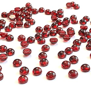 May include: A scattering of small, round, dark red glass beads. The beads are domed on one side and flat on the other, with a glossy finish. They are scattered across a white surface, creating a visually appealing display.