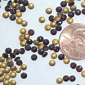 Puede incluir: Una imagen de primer plano de una colección de pequeñas cuentas redondas de color rojo y dorado. Las cuentas están esparcidas sobre una superficie blanca. Un centavo de euro se incluye en la imagen para la escala.