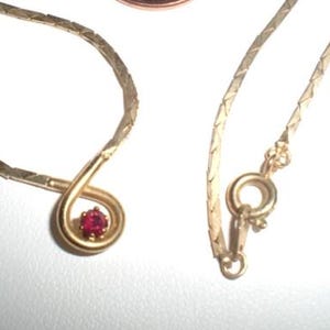 May include: A gold-toned necklace with a small, red gemstone pendant. The necklace is next to a United States one-cent coin for size comparison. The chain has a herringbone design and a clasp closure.