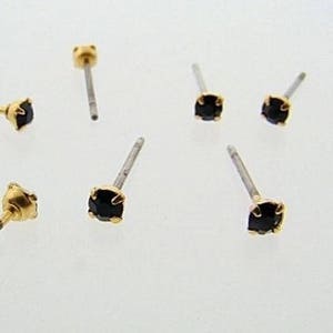 May include: A collection of small stud earrings with black gemstones. Each earring features a gold-colored setting and a silver-colored post. The earrings are scattered against a white background, showcasing their design.