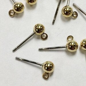 May include: A close-up of ten gold earring findings with a small ball at the end of each post. The findings are on a white surface.