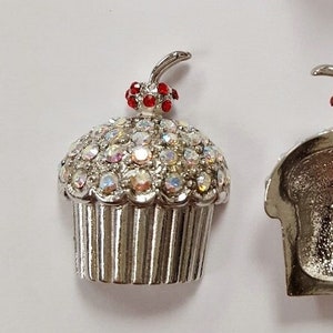 May include: Silver cupcake charm with a red cherry on top. The cupcake is covered in iridescent rhinestones. The charm is shown next to a US penny for size comparison.