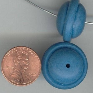 May include: Two blue ceramic beads with a hole in the centre. The beads are round and have a textured surface. One bead has a raised ring around the centre. The beads are about the size of a UK penny.