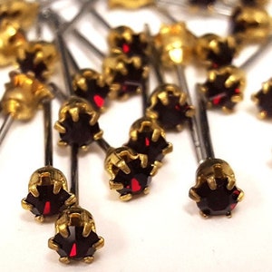 Puede incluir: Primer plano de un grupo de tachuelas de strass rojas con engastes dorados. Las tachuelas están unidas a postes plateados.