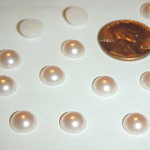 May include: A collection of white pearl-like buttons, some are round and some are slightly oval shaped, arranged on a white surface. A penny is included for size comparison.