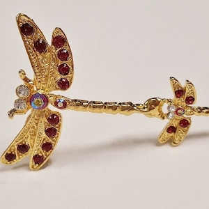 May include: Gold-tone dragonfly brooch with red and clear rhinestones. The dragonfly has a detailed design and is attached to a long, thin chain.