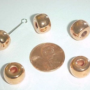 May include: Five shiny copper-colored beads with a hole in the center. The beads are round and smooth. They are arranged in a semi-circle on a white surface.