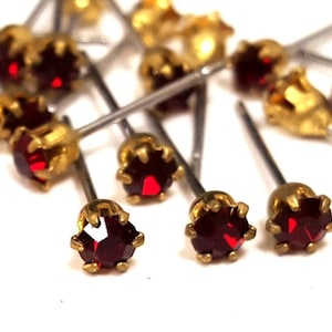 Puede incluir: Primer plano de un grupo de alfileres de strass rojos con engastes dorados. Los alfileres están unidos a postes de metal plateado.