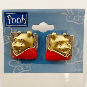 Puede incluir: Un par de pendientes de Winnie de Pooh en tono dorado. Cada pendiente tiene un diseño cuadrado con un revestimiento de esmalte rojo. La imagen de Winnie de Pooh está en el centro de cada pendiente. Los pendientes están en una tarjeta azul y blanca con la palabra "Pooh" impresa en ella. © Disney.
