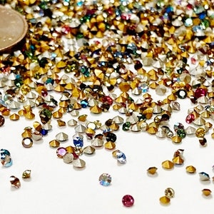 May include: A large assortment of colorful rhinestones, some gold, some silver, and some with iridescent finishes. The rhinestones are scattered on a white surface. A US penny is included for scale.
