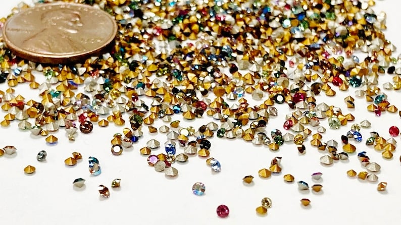 May include: A large assortment of colorful rhinestones, some gold, some silver, and some with iridescent finishes. The rhinestones are scattered on a white surface. A US penny is included for scale.