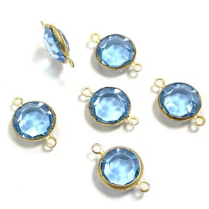 May include: Six light blue gemstone charms with gold-colored metal settings. The charms are round with faceted surfaces, and each has two small loops for attaching to jewelry. One charm is oval-shaped.