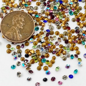 May include: A close-up of a large assortment of colorful rhinestones, with a gold-colored penny featuring the text "In God We Trust" and "Liberty" in the background.