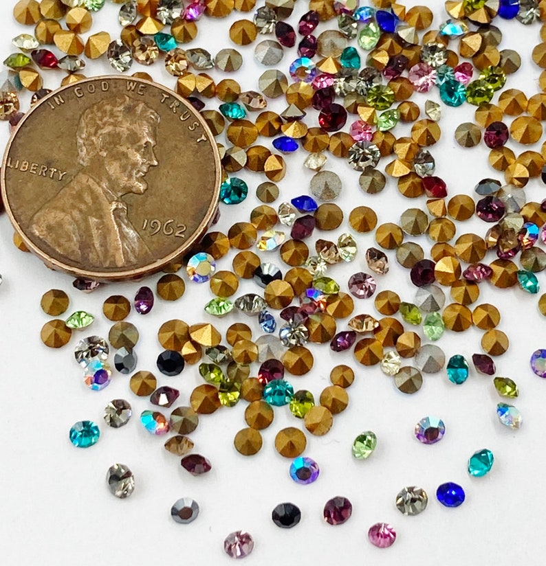 May include: A close-up of a large assortment of colorful rhinestones, with a gold-colored penny featuring the text "In God We Trust" and "Liberty" in the background.