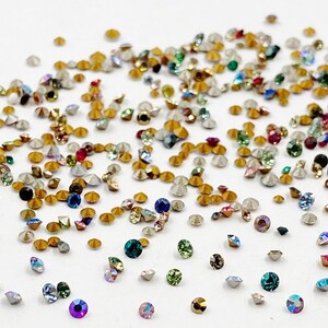 May include: Assortment of small, colorful, faceted rhinestones with gold settings. The rhinestones are scattered on a white surface.