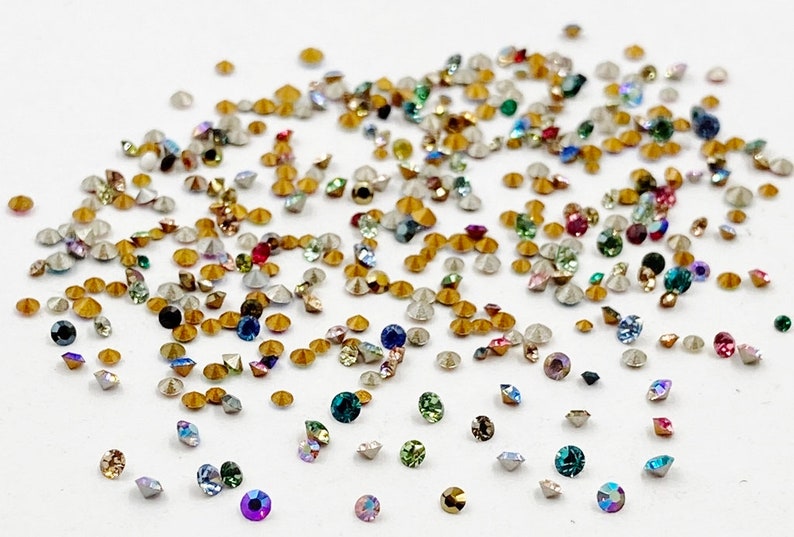 May include: Assortment of small, colorful, faceted rhinestones with gold settings. The rhinestones are scattered on a white surface.