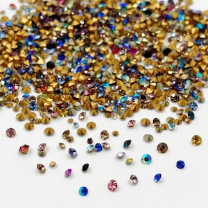 May include: Assortment of colorful rhinestones in various shapes and sizes, including round, square, and star-shaped. The rhinestones are scattered on a white surface.