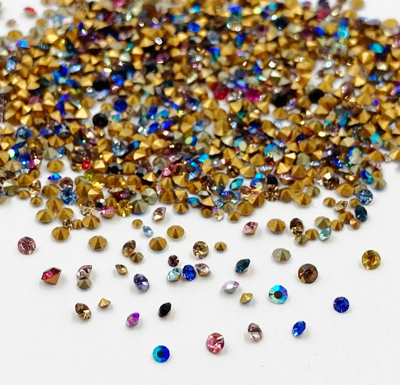 May include: Assortment of colorful rhinestones in various shapes and sizes, including round, square, and star-shaped. The rhinestones are scattered on a white surface.
