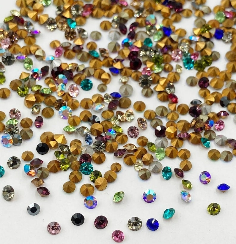 May include: Assortment of colorful glass rhinestones in various shapes and sizes, including round, square, and star shapes. The rhinestones are scattered on a white background.