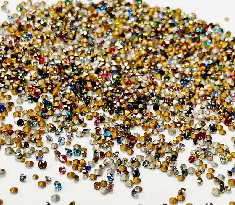 May include: A large assortment of colorful rhinestones in various sizes and shapes. The rhinestones are scattered on a white surface.