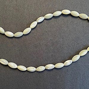 May include: A necklace of oval-shaped, pale green beads. The beads are strung together on a thin, clear string, creating a delicate and elegant design. The beads have a subtle sheen, and the necklace is displayed against a dark background.