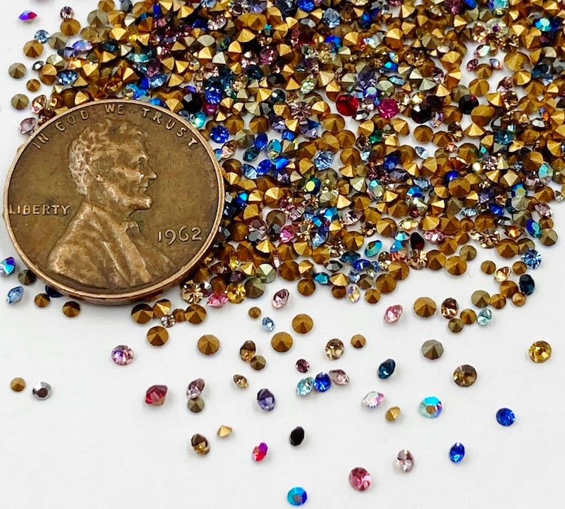May include: A close-up image of a variety of colorful rhinestones and gold-toned metal beads. The beads are scattered on a white surface. A US penny coin with the year 1962 is visible in the image.