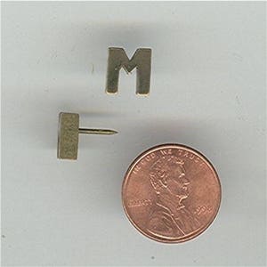 May include: A gold-colored letter M pin with a push pin attached to the back. The pin is next to a US penny with the year 1998.