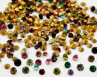 May include: Assortment of colorful rhinestones in various shapes and sizes, including round, square, and star-shaped. The rhinestones are scattered on a white surface.