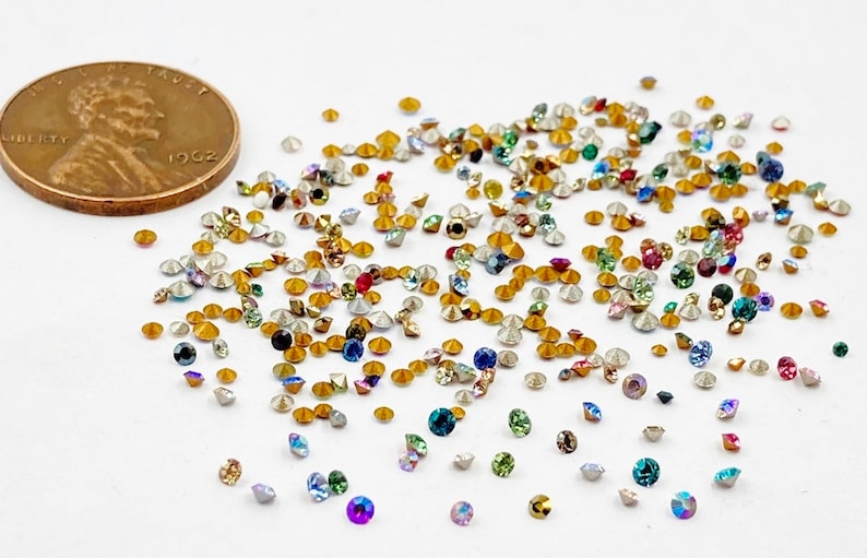 May include: A large assortment of small, colorful, iridescent rhinestones. The rhinestones are scattered on a white surface. A US penny is included for scale.