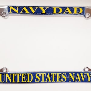 May include: A chrome license plate frame with blue and yellow text. The top bar reads "NAVY DAD" and the bottom bar reads "UNITED STATES NAVY" with an anchor emblem on each end. The frame is designed for a standard US license plate.