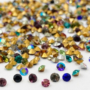 May include: Assortment of colorful rhinestones in various shapes and sizes, including gold, silver, green, blue, purple, and black. The rhinestones are scattered on a white surface.