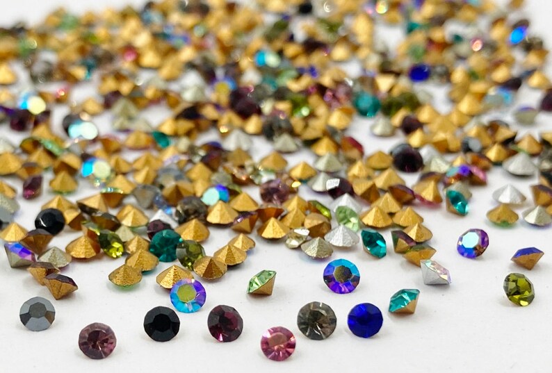 May include: Assortment of colorful rhinestones in various shapes and sizes, including gold, silver, green, blue, purple, and black. The rhinestones are scattered on a white surface.
