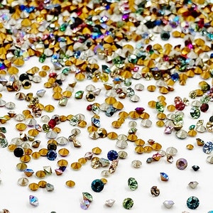 May include: Assortment of colorful rhinestones in various sizes and shapes, including gold, silver, clear, and iridescent colors. The rhinestones are scattered on a white background.
