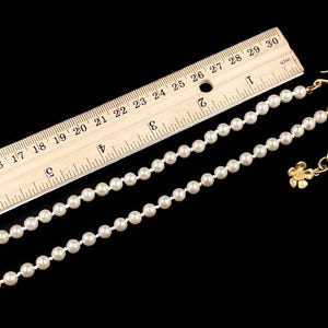 May include: A white pearl necklace with a gold chain and a small flower charm. The necklace is measured against a ruler, showing it is approximately 18 inches long.