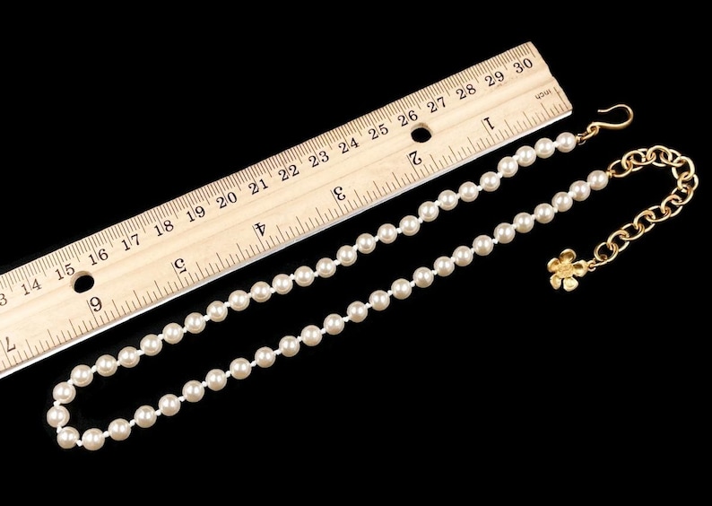 May include: A white pearl necklace with a gold chain and a small flower charm. The necklace is measured against a ruler, showing it is approximately 18 inches long.