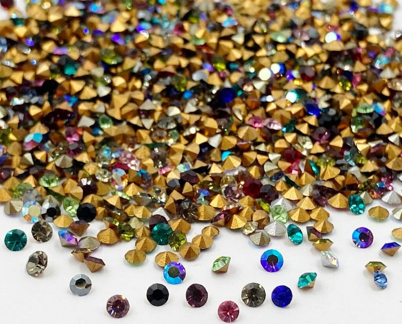 May include: A large pile of colorful rhinestones with gold settings. The rhinestones are scattered on a white surface.