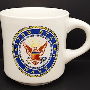 Pode incluir: Uma caneca de cerâmica branca com o emblema da Marinha dos Estados Unidos impresso na frente. O logótipo circular inclui uma borda de corda amarela, um fundo azul e um gráfico central de uma águia, escudo e âncora com o texto UNITED STATES NAVY.
