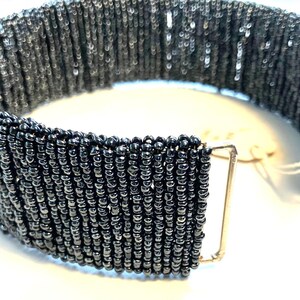 May include: A wide, dark gray beaded bracelet with a rectangular clasp. The beads are small and tightly strung together, creating a textured surface. The clasp is a light gold color. The bracelet is likely for the wrist.