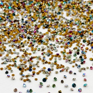 May include: A large assortment of colorful rhinestones, with a variety of shapes and sizes, scattered on a white surface.